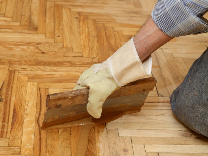 Bodenleger Hand mit Handschuh bringt Holzparkett mit Spachtel auf