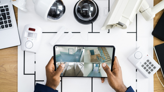 Förderungen und Finanzierungen Person hält Tablet mit Überwachungskamera-Feeds über einem Tisch mit Sicherheitsausrüstung