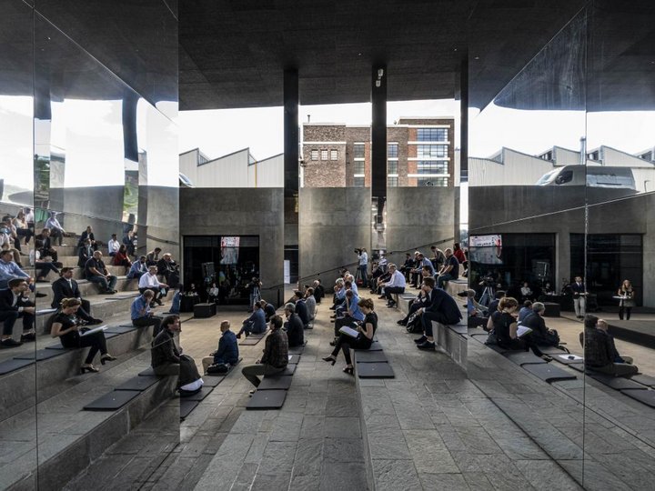 Zusammenarbeit & Netzwerke Menschen sitzen in einem modernen, offenen Veranstaltungsraum mit Spiegelwänden