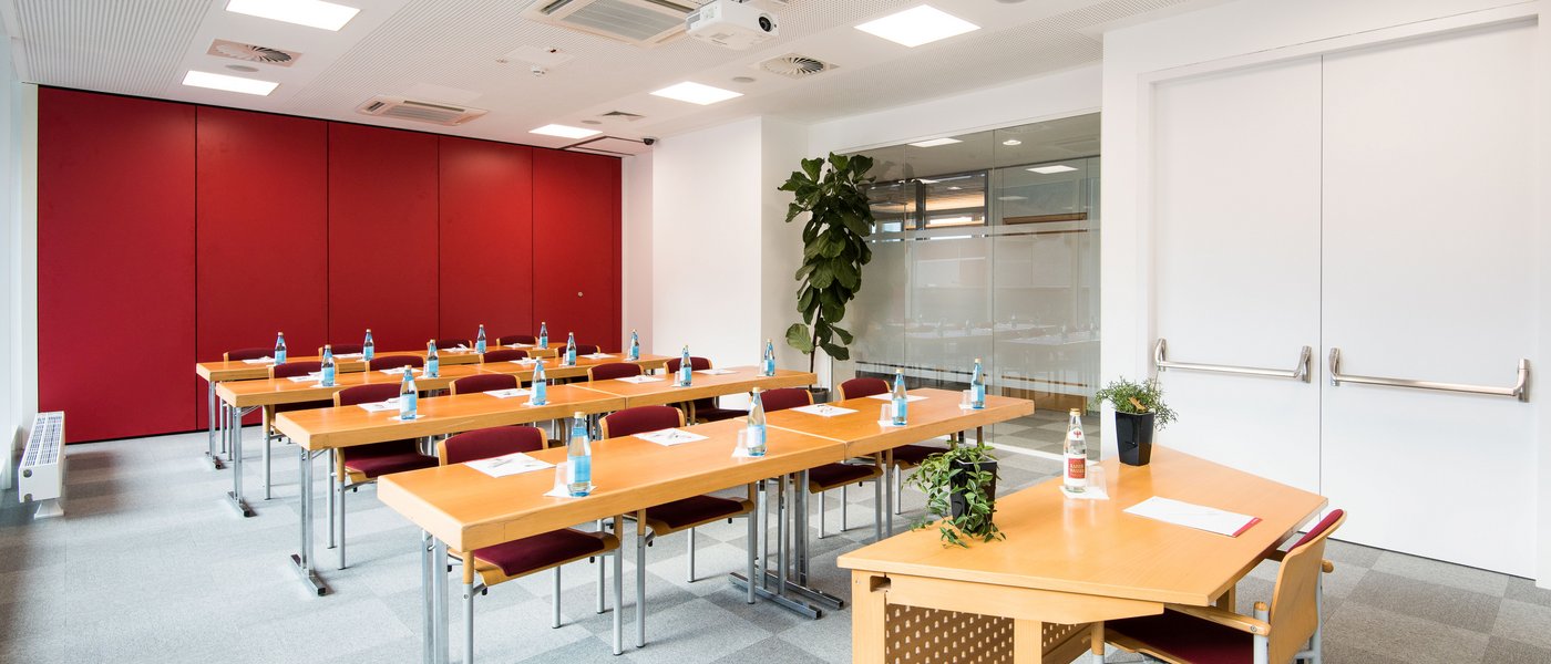 Vermietung Seminarräume Heller Schulungsraum mit Holztischen, roten Stühlen und Wasserflaschen