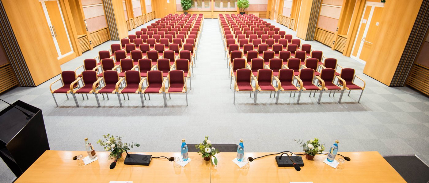 Vermietung Seminarräume Leerer Vortragssaal mit Reihen von roten Stühlen und Tisch mit Mikrofonen