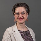 Schönheitspfleger Porträt einer lächelnden Frau mit Brille und beiger Jacke vor grauem Hintergrund