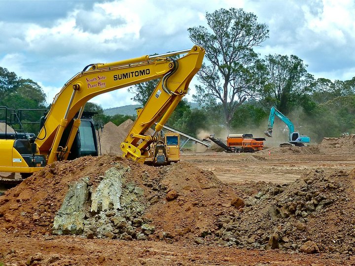 Tiefbauunternehmer Baustelle mit gelbem und blauem Bagger, Erde und Bäumen im Hintergrund
