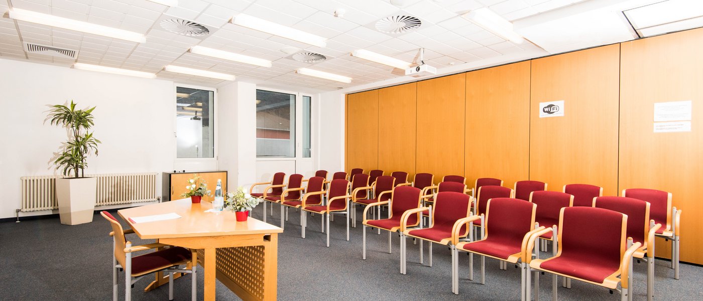 Vermietung Seminarräume Leerer Konferenzraum mit Reihen roter Stühle, Holztisch und Fenster