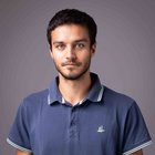 Weiterbildung Porträt eines jungen Mannes mit dunklem Haar und blauem Poloshirt vor grauem Hintergrund