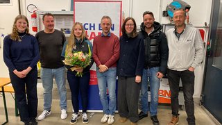 News Sieben Personen stehen in einer Werkstatt vor einem Banner, eine hält Blumen