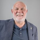 Tapezierer - Raumausstatter Älterer Mann mit grauem Bart und blauem Sakko vor grauem Hintergrund