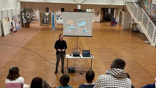 Junghandwerker Frau hält Präsentation vor kleiner Gruppe in großem, hellem Raum mit Stufen