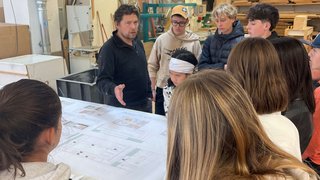 Junghandwerker Lehrer erklärt Schülern technische Zeichnungen in einer Werkstatt mit Holz und Maschinen