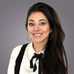 Das lvh-Team Lächelnde Frau mit langen schwarzen Haaren und goldenen Ohrringen vor grauem Hintergrund