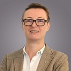 Funktionäre Porträt einer Person mit Brille und braunem Blazer vor grauem Hintergrund