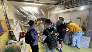 Junghandwerker Gruppe Jugendlicher hört einem Maler in Werkstatt mit Farbeimern und Werkzeug zu