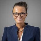 Baugewerbe Porträt einer Frau mit Brille, blauem Blazer und Perlenkette vor grauem Hintergrund
