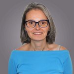 Das lvh-Team Frau mit grauen Haaren und blauer Brille lächelt in blauem Oberteil vor grauem Hintergrund