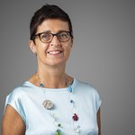 Das lvh-Team Porträt einer lächelnden Frau mit kurzen Haaren, Brille und türkisem Oberteil vor grauem Hintergrund