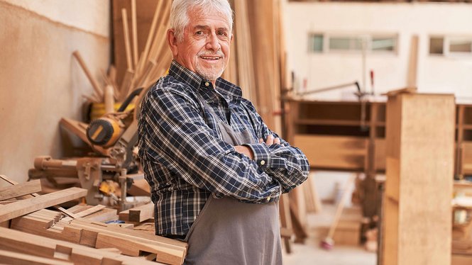 Home Älterer Handwerker in Werkstatt mit Holzbrettern und Werkbank