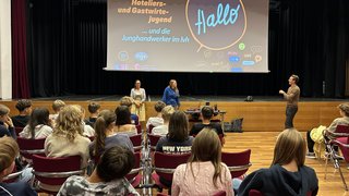 News Jugendliche sitzen in einem Saal vor einer Bühne mit Präsentation und sprechen mit drei Erwachsenen