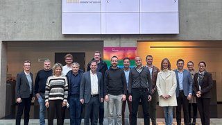 News Gruppenfoto von 15 Personen in einem modernen Gebäude mit digitaler Anzeigetafel