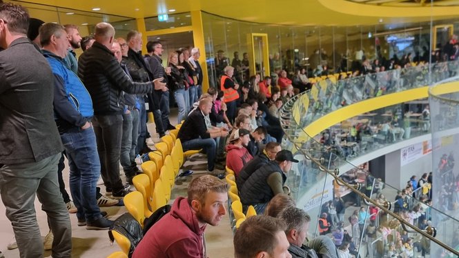 Home Zuschauer verfolgen ein Sportereignis von gelben Tribünensitzen hinter einer Glasbrüstung