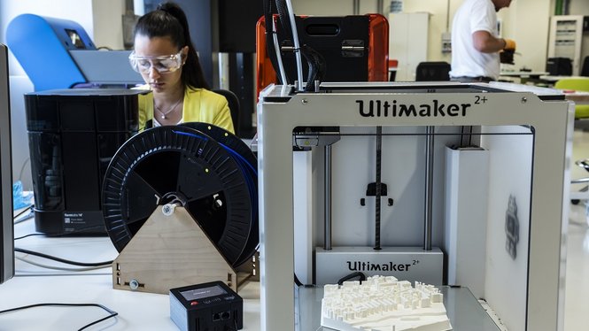 Home Frau mit Schutzbrille arbeitet neben einem Ultimaker-3D-Drucker, im Hintergrund ein Mann