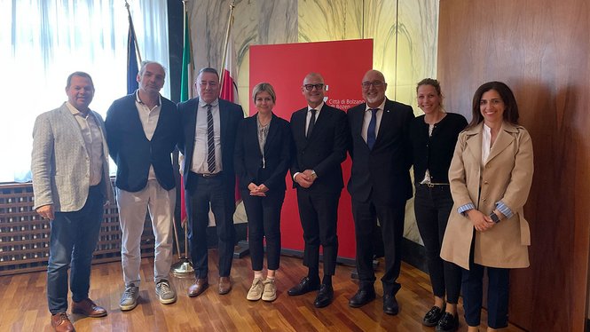 Home Gruppenfoto von acht Personen vor Fahnen und rotem Stadt Bozen Schild