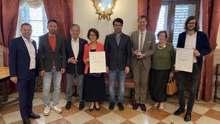 News Neun Personen stehen in festlicher Umgebung mit Urkunden und Medaille