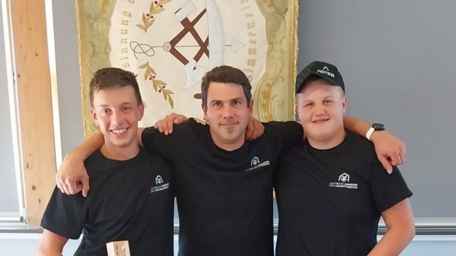 Home Drei Männer in schwarzen T-Shirts posieren vor einem Wappen an der Wand