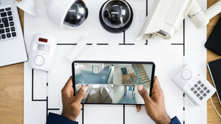 News Person hält Tablet mit Überwachungskamera-Feeds über einem Tisch mit Sicherheitsausrüstung