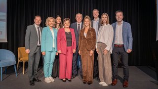 News Neun Personen in Businesskleidung posieren vor schwarzem Vorhang auf Bühne