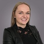 Das lvh-Team Porträt einer lächelnden jungen Frau mit schwarzer Spitzenbluse und Lederjacke vor grauem Hintergrund