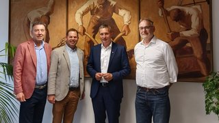 News Vier Männer stehen vor einem dreiteiligen Gemälde mit arbeitenden Figuren