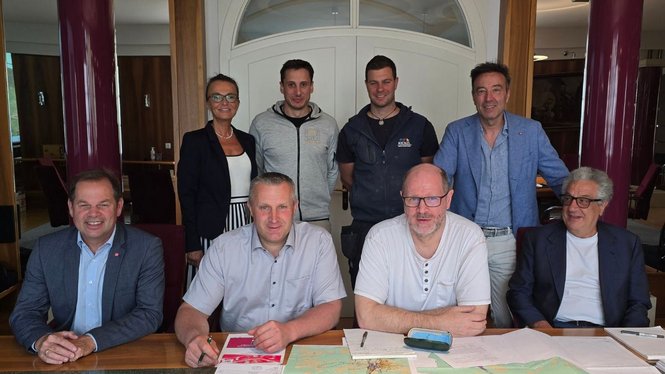 Home Acht Personen in einem Raum, vier sitzen an einem Tisch mit Papieren, vier stehen dahinter