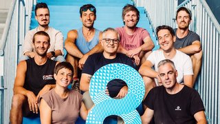 News Gruppe von neun Personen sitzt auf blauen Treppen, hält große blaue Zahl 8