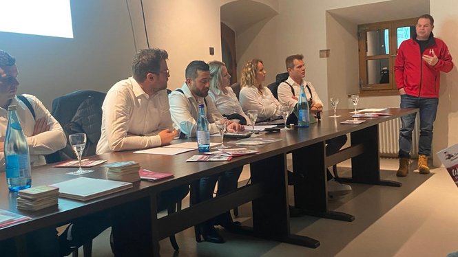 Home Personen sitzen an einem Tisch und hören einem stehend Vortragenden in rotem Jacke zu