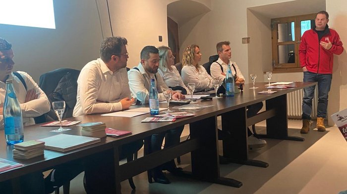 Tag des Kachelofens am 20. Oktober Personen sitzen an einem Tisch und hören einem stehend Vortragenden in rotem Jacke zu