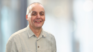 News Uomo con camicia grigia che sorride davanti a sfondo sfocato