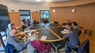 News Nove persone sedute intorno a un tavolo rotondo in una sala riunioni.