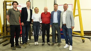 News Sei persone in piedi in un capannone davanti a macchinari e striscioni.