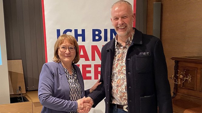 Home Zwei Personen geben sich die Hand und lächeln vor einem Plakat