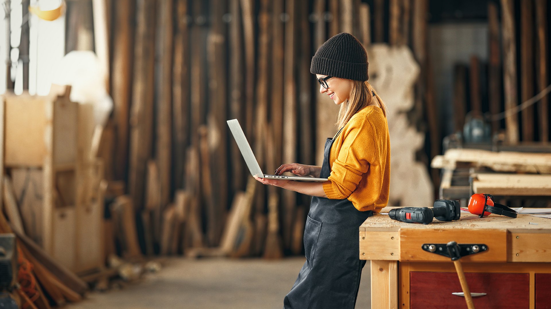 Home Frau mit Mütze in Werkstatt arbeitet am Laptop neben Holz und Werkzeug