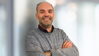 News Uomo sorridente con maglione grigio e braccia incrociate su sfondo chiaro