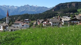 News Bergdorf mit Kirche und grünen Wiesen vor Alpenkulisse bei klarem Himmel