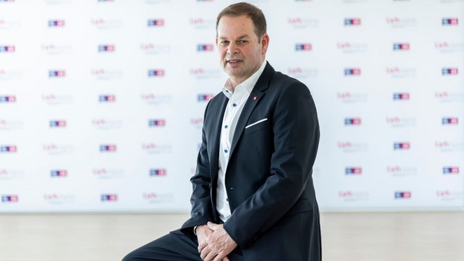 Home Mann im dunklen Anzug sitzt vor einer Wand mit wiederkehrenden Logos