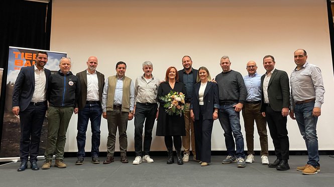 Home Gruppe von elf Personen steht vor weißer Leinwand auf Bühne, eine Frau hält Blumenstrauß