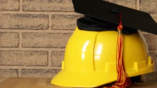 News Casco da cantiere giallo con tocco da laurea nero e nappina rossa davanti a muro di mattoni
