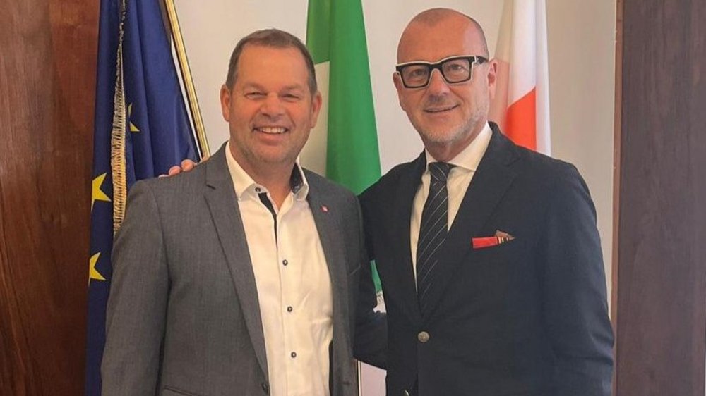 Home Zwei Männer in Anzügen lächeln vor drei Flaggen in einem Innenraum