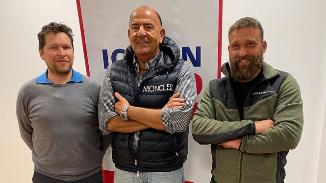 Home Drei Männer stehen lächelnd vor einer weißen Wand mit rotem und blauem Banner