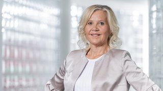 News Ältere Frau mit hellblondem Haar in silberner Jacke lächelnd vor unscharfem Hintergrund