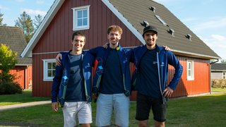 News Drei junge Männer mit blauen Jacken posieren vor einem roten Haus im Freien