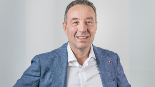 News Porträt eines lächelnden Mannes im blauen Sakko vor neutralem Hintergrund